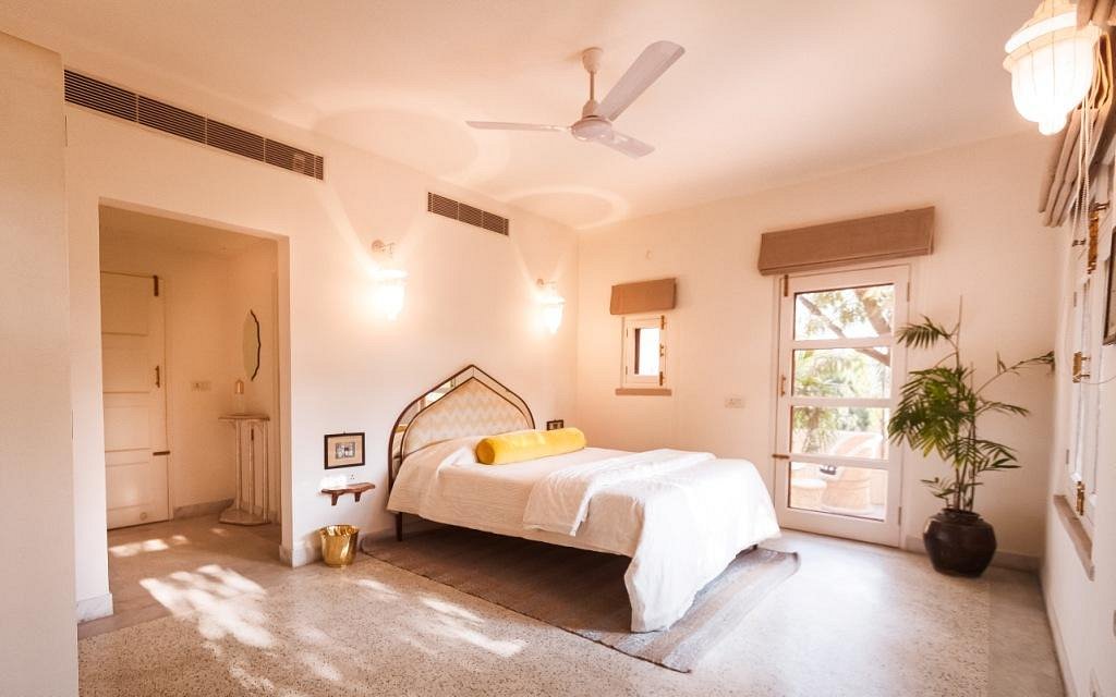 12 BEST Boutique Hotels in Jaipur 33 A bright, airy bedroom inspired by boutique hotels in Jaipur features white walls, a double bed with a decorative headboard, white bedding accented with yellow, two windows, a balcony door, potted plant, and stylish wall lights.