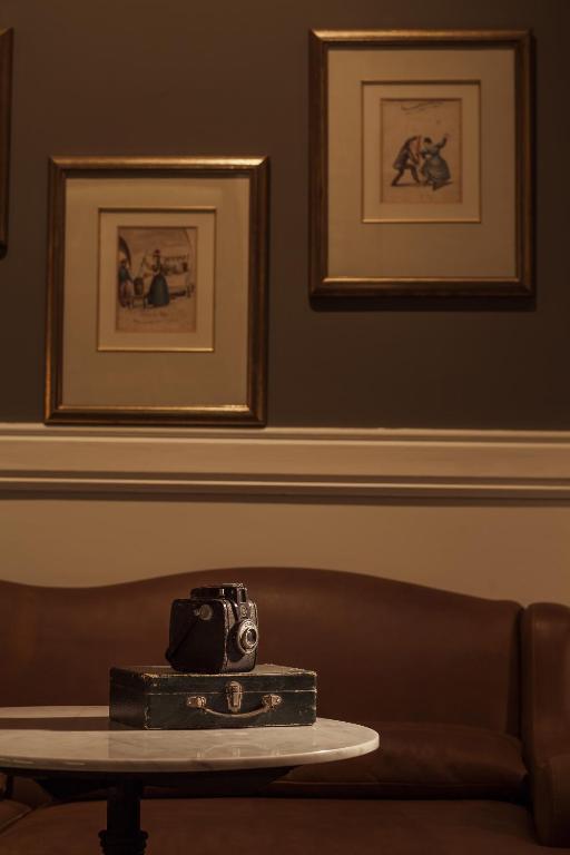 9 Unique Boutique Hotels in Lima 29 A vintage camera sits on a closed suitcase atop a round marble table, evoking the eclectic charm of boutique hotels in Lima, with a brown leather couch and three framed artworks adorning the taupe wall behind.