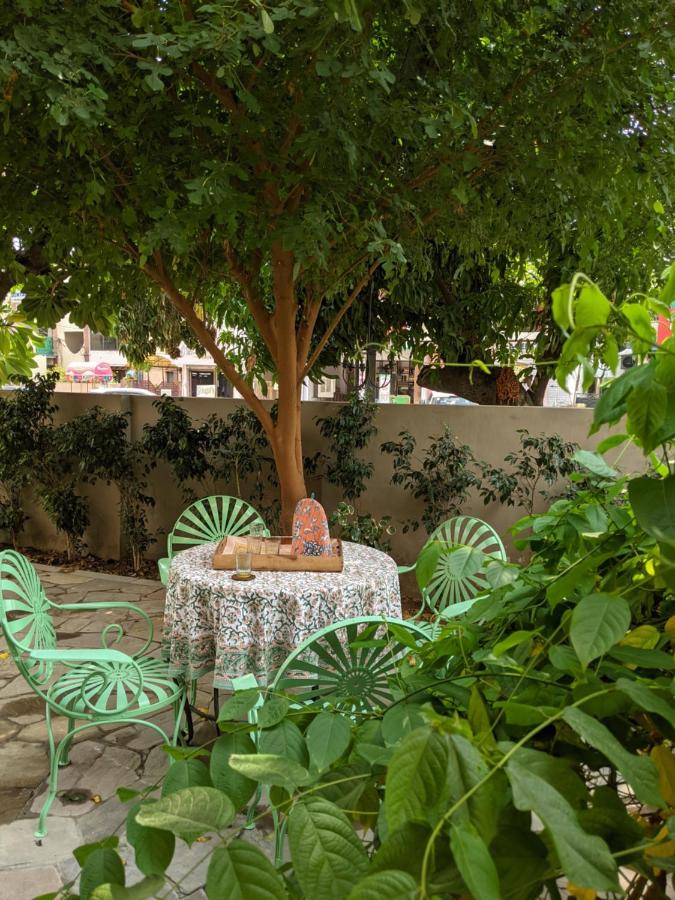 12 BEST Boutique Hotels in Jaipur 16 A round table with a floral tablecloth and mint green chairs sits under a leafy tree in a garden—a scene reminiscent of boutique hotels in Jaipur. Wooden boxes, a ceramic pitcher, and a glass rest on the table, surrounded by lush greenery.