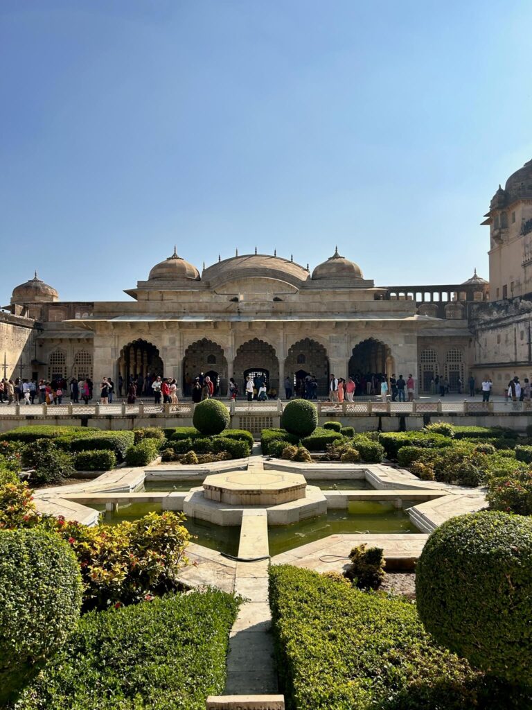 12 BEST Boutique Hotels in Jaipur 52 A Mughal-style palace with domes and arched columns stands behind a manicured garden with trimmed bushes, a central fountain, and a small pond under a clear blue sky—reminiscent of boutique hotels in Jaipur. Visitors stroll near the palace entrance.