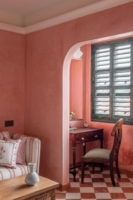 12 BEST Boutique Hotels in Jaipur 21 A cozy room inspired by boutique hotels in Jaipur features warm pink walls, a striped sofa with patterned pillows, a wooden desk and chair by green-shuttered windows, and checkered tile floors for an inviting touch.