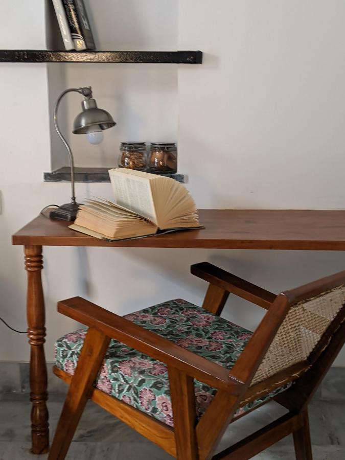 12 BEST Boutique Hotels in Jaipur 18 A wooden desk with an open book, a vintage-style lamp, two jars, and a floral-cushioned chair creates a cozy corner reminiscent of boutique hotels in Jaipur. A shelf with books is mounted on the white wall above the desk.