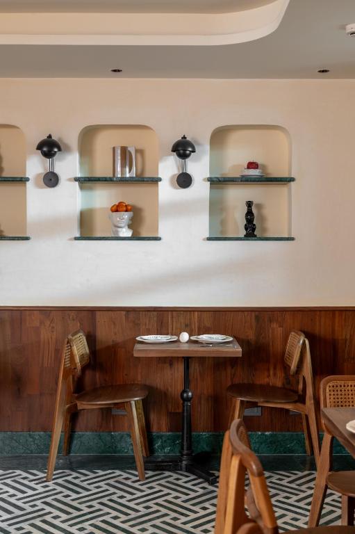 12 BEST Boutique Hotels in Jaipur 22 A small restaurant table set for two with wooden chairs, against a wall with built-in shelves displaying decor items and lit by black wall sconces—reminiscent of the chic interiors found in boutique hotels in Jaipur. The floor has a geometric black-and-white pattern.