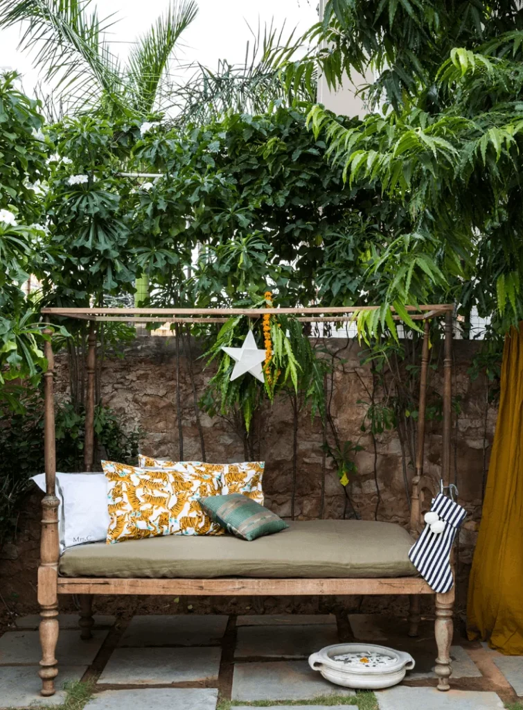 12 BEST Boutique Hotels in Jaipur 7 A rustic wooden daybed with patterned cushions sits outdoors beneath lush green trees at one of the charming boutique hotels in Jaipur. A white star decoration hangs above, with a striped bag and decorative plate nearby, creating a cozy garden nook.