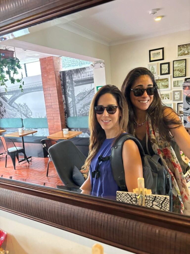 9 Unique Boutique Hotels in Lima 21 Irene and friend Karla wearing sunglasses and backpacks smile while posing together for a mirror selfie in a cozy café, reminiscent of the stylish lounges found in boutique hotels in Lima, with framed photos and art on the walls.