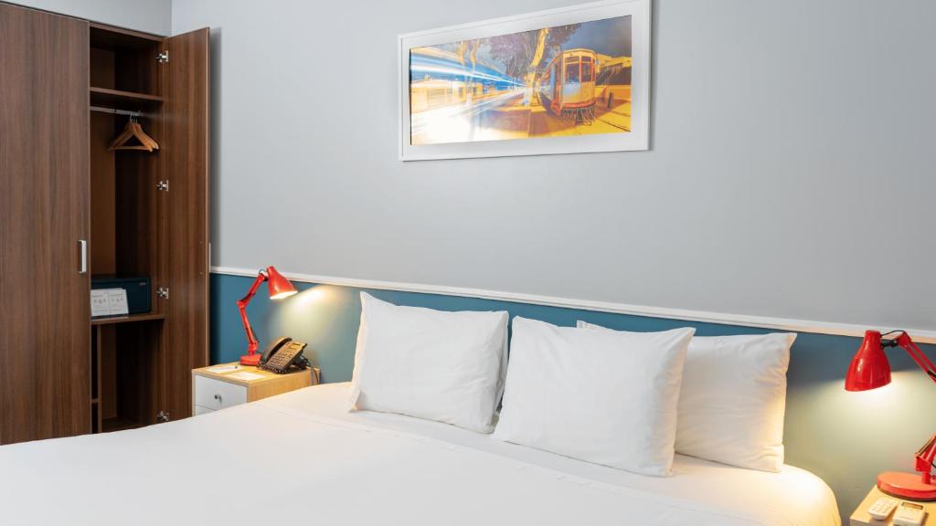 9 Unique Boutique Hotels in Lima 42 A neatly made bed with three white pillows, two red desk lamps on bedside tables, an open wardrobe, and a wall-mounted phone reflects the stylish comfort found in boutique hotels in Lima, with a framed street scene above the bed.