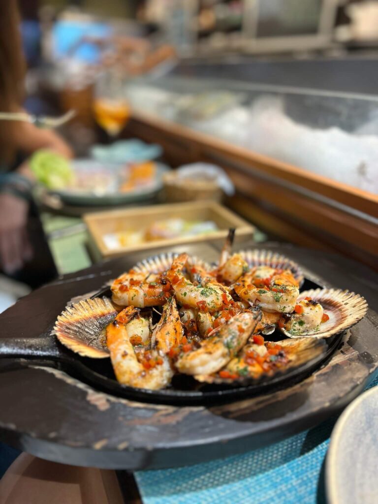 A close-up of grilled seafood, including shrimp and shellfish, garnished with herbs and roe, served on a black plate at a restaurant counter in one of the boutique hotels in Lima, with other dishes and drinks in the background.