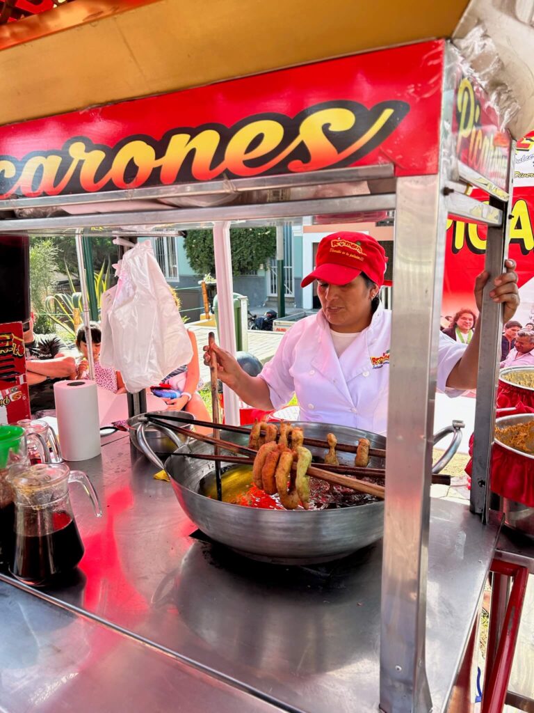 A woman in a white uniform and red cap fries churros in a large pan at a street food stall—an authentic taste of Lima you might discover near boutique hotels in Lima, with bottles of syrup and a cup beside her under a yellow and red sign.