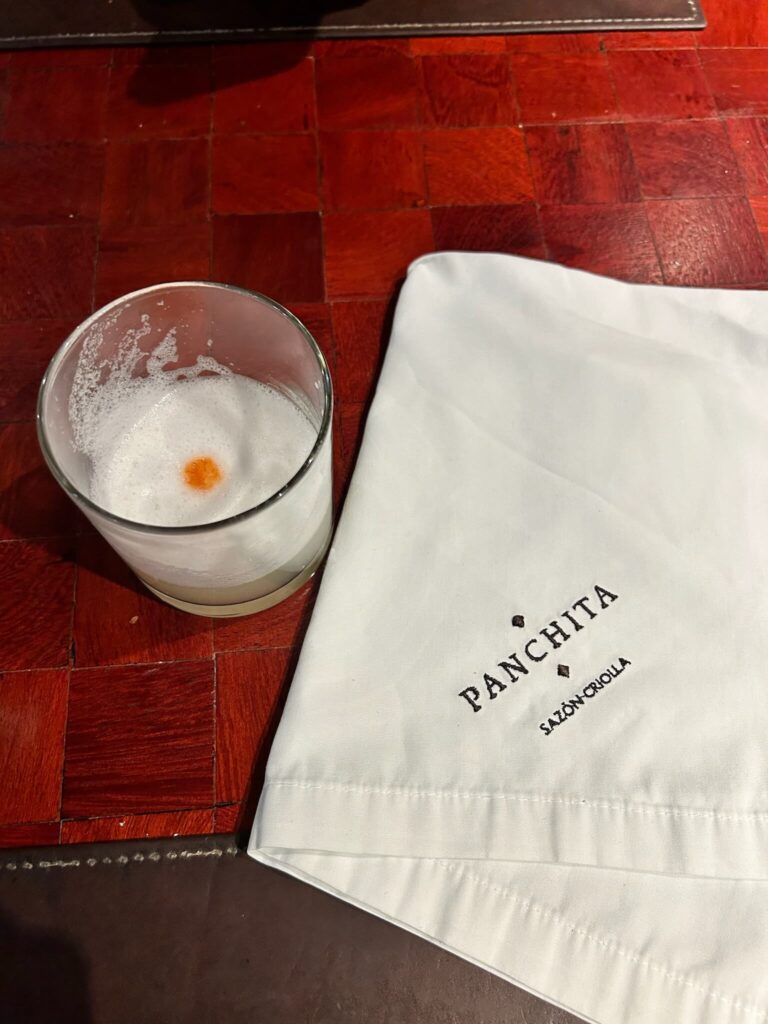 A glass of pisco sour with a frothy top and a drop of bitters sits on a red checkered table in one of the charming boutique hotels in Lima, next to a white napkin embroidered with the words Panchita Sazón Criolla.