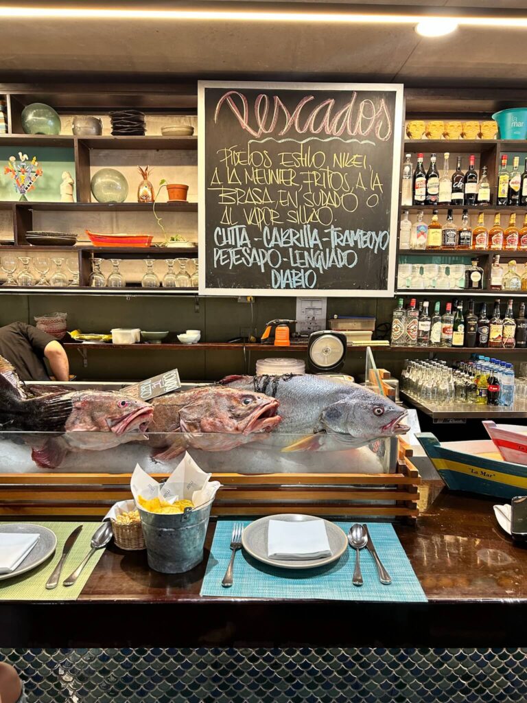 A restaurant bar in one of the boutique hotels in Lima displays four whole fresh fish on ice beneath a chalkboard menu listing various types. Shelves behind the bar hold bottles, glasses, and plates; a table is set with food and utensils in the foreground.