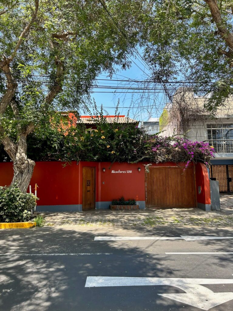 9 Unique Boutique Hotels in Lima 2 A red-walled house with a wooden door and garage, partially shaded by tall trees and flowering plants, sits on a sunny street with a crosswalk in front—reminiscent of the charm found at boutique hotels in Lima. The address Alcanfores 1281 is visible on the wall.