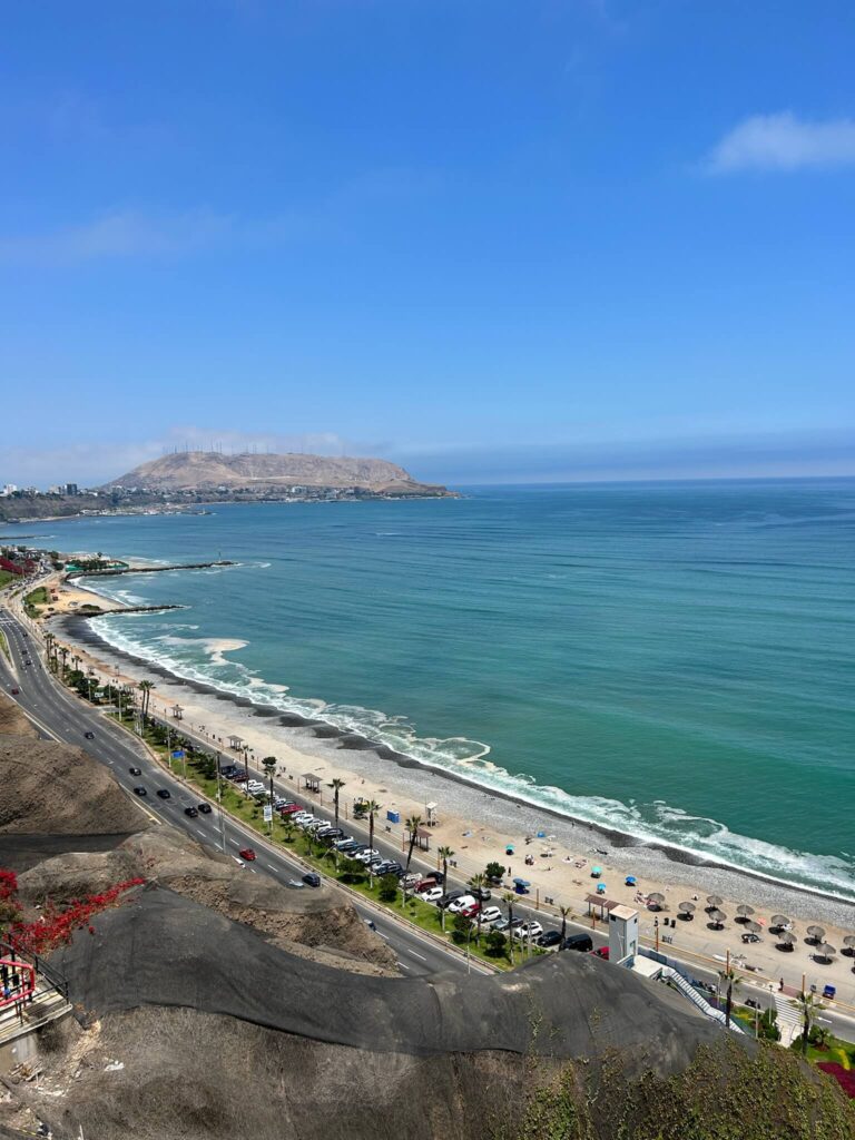 9 Unique Boutique Hotels in Lima 1 A scenic view of a coastal road running alongside a sandy beach with turquoise waves, palm trees, and cars parked near the shore under a bright blue sky with distant hills—perfect for visitors staying at boutique hotels in Lima.