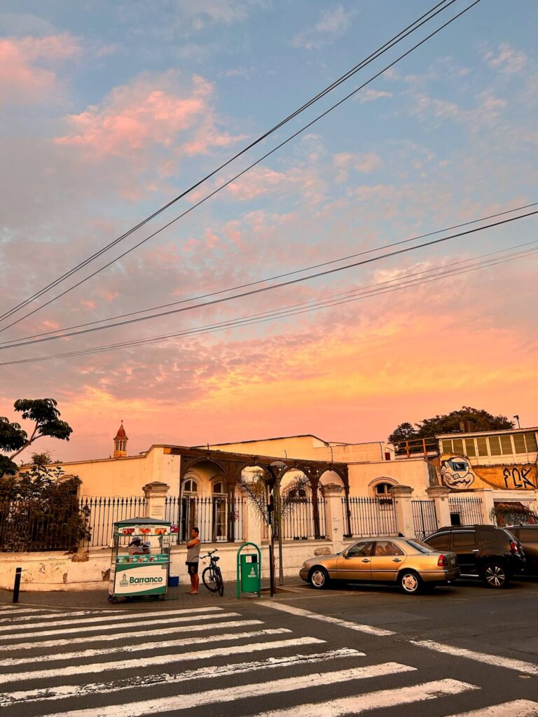 9 Unique Boutique Hotels in Lima 3 A street scene at sunset shows pastel clouds in the sky, a crosswalk, parked cars, a mailbox, and a small street vendor cart near boutique hotels in Lima with white-walled buildings, arched windows, and a fence.