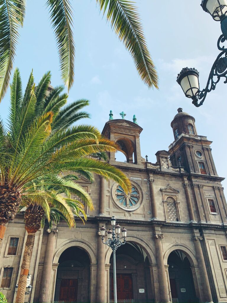 Unique Hotels in Spain 14 A historic stone cathedral with arched windows and towers, partially framed by palm tree leaves and an ornate street lamp, under a bright blue sky—reminiscent of the charm found near adults only hotels in Gran Canaria.