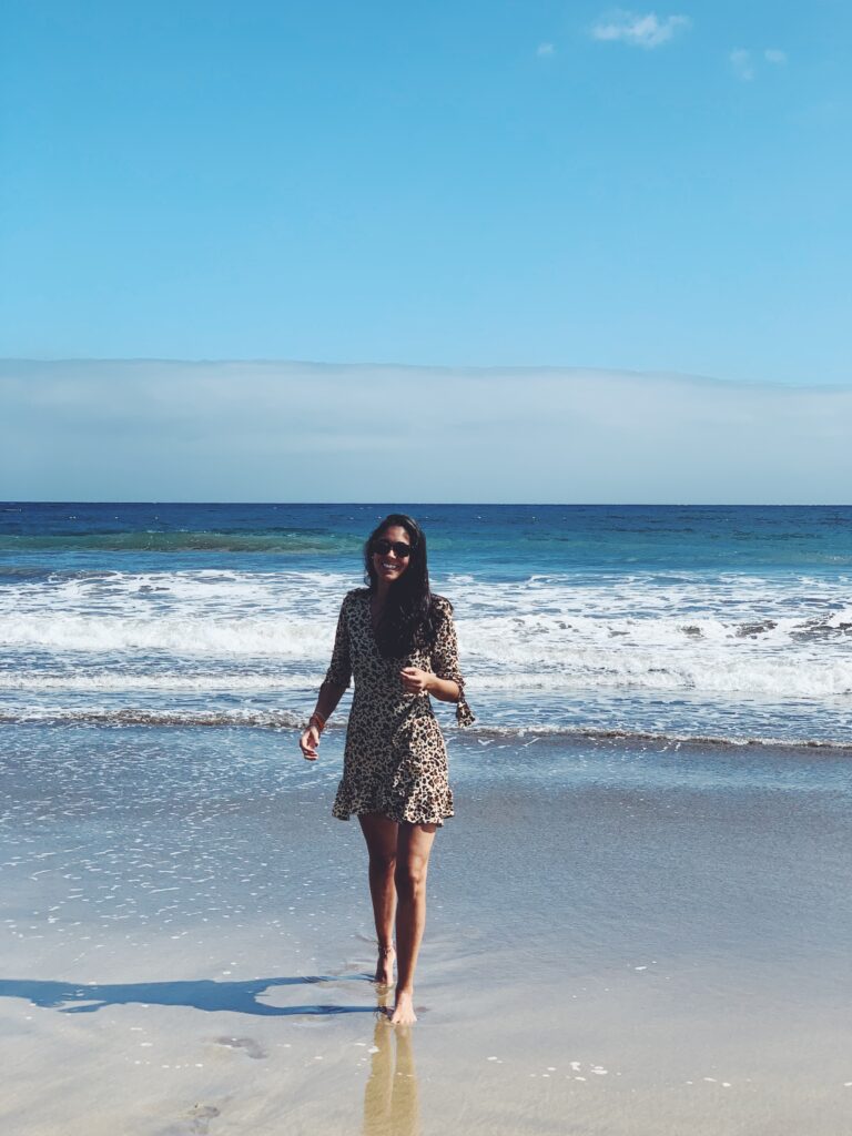 Unique Hotels in Spain 13 A woman wearing a patterned dress and sunglasses stands barefoot at the shoreline, smiling, with waves and a blue sky in the background—evoking carefree moments at adults only hotels in Gran Canaria.