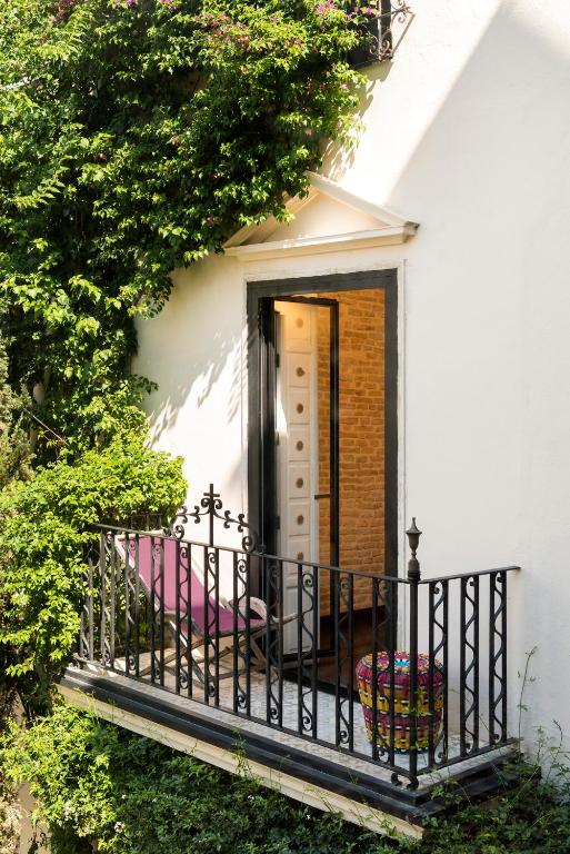 A small balcony with ornate black railings, a pink lounge chair, and a colorful basket graces a white building draped in lush ivy—evoking the charm of boutique hotels in Seville. The open door reveals a warmly lit brick interior.