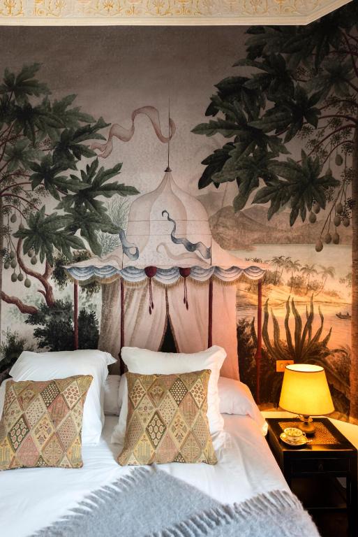 A bed with patterned pillows and a cozy blanket sits in front of a wall mural depicting a tent, trees, and a scenic landscape—evoking the unique charm found in boutique hotels in Seville; a lit lamp and small items rest on the bedside table.