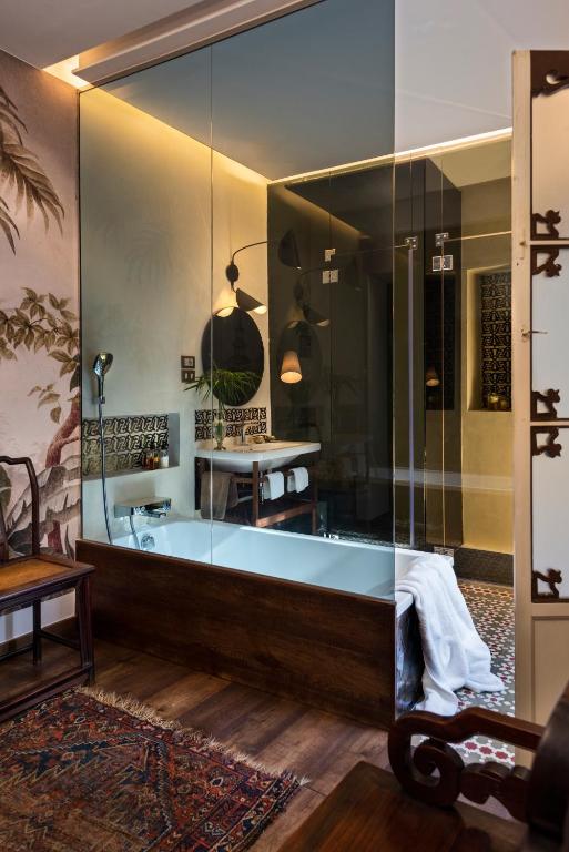 A stylish bathroom reminiscent of boutique hotels in Seville, with a glass wall, wooden floors, a white bathtub, modern sink, round mirror, wall lamps, patterned tiles, and decorative wallpaper. A colorful rug and wooden furniture add warmth.