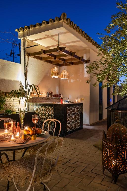 A cozy outdoor patio at night, reminiscent of boutique hotels in Seville, features a small bar with patterned tiles, hanging lights, candles, a table set with wine glasses and fruit, a fringed chair, all surrounded by lush greenery.