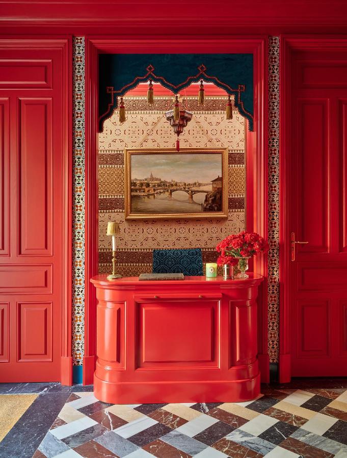A vibrant red entryway, reminiscent of the stylish interiors found in boutique hotels in Seville, features ornate paneling, a matching red console table, flowers, patterned walls, and a framed landscape painting above, set on a geometric black and white tile floor.