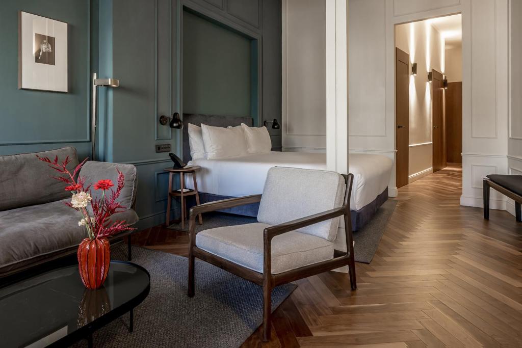 Modern hotel room with a double bed, gray sofa, armchair, wooden floors, art on the wall, and a red vase with flowers on a black coffee table. Bright, well-lit space with a hallway in the background—evoking the stylish elegance found in boutique hotels in Seville.