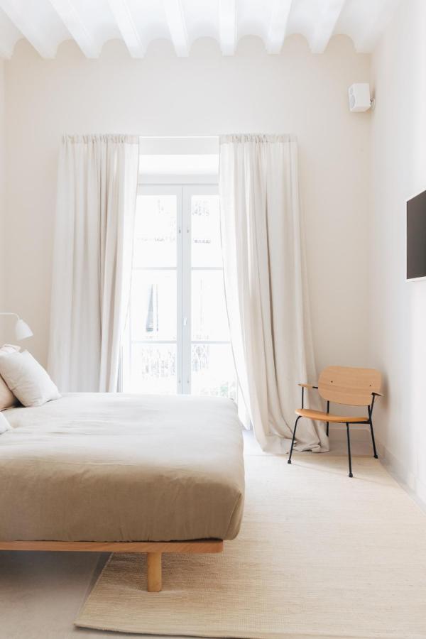 Minimalist bedroom with a beige bed, light wooden chair, cream curtains, large window, white walls, and a wall-mounted TV. The bright, airy space echoes the elegant style found in boutique hotels in Seville.