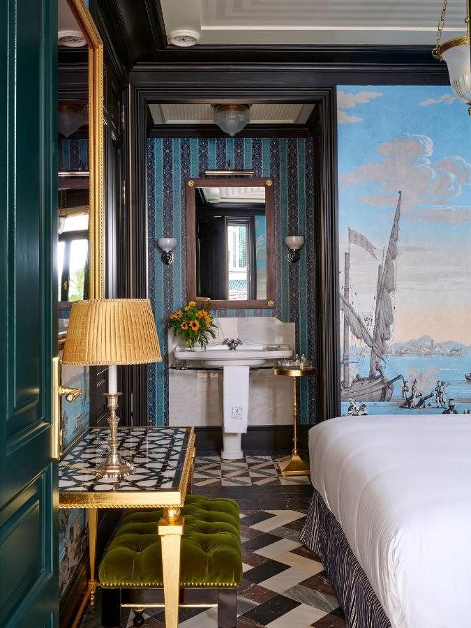 A stylish hotel room reminiscent of the boutique hotels in Seville, with patterned floors, a green velvet bench, a lamp on a tiled desk, a bed with white linens, a sink with a mirror, and a wall mural featuring a sailboat and ocean scene.