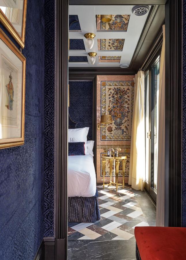 Luxurious bedroom with deep blue textured walls, framed art, patterned black and white floor, ornate gold-accented decor, and a sunlit bed next to floor-to-ceiling windows with light curtains—evoking the sophisticated charm often found in boutique hotels in Seville.