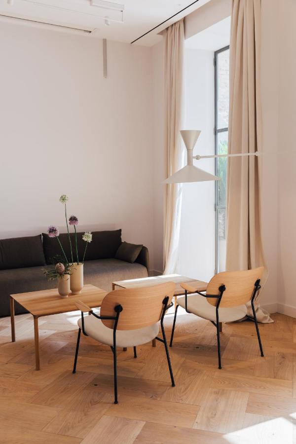 A modern, minimalist living room inspired by boutique hotels in Seville, featuring light wood floors, a black sofa, wood chairs, a wooden coffee table with vases of flowers, tall beige curtains, a large window, and a white pendant lamp.
