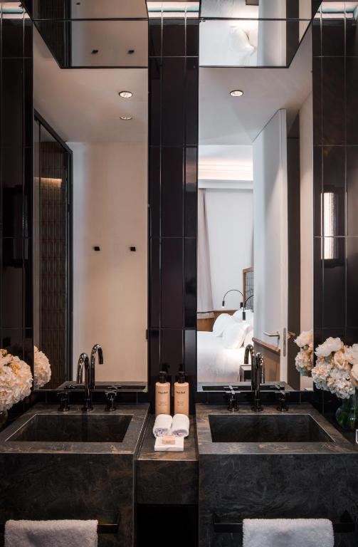 Modern bathroom with two black sinks, black fixtures, large mirrors, white towels, flower arrangements, and toiletries—evoking the refined elegance found in boutique hotels in Seville. A bedroom with white bedding is visible through an open door in the background.