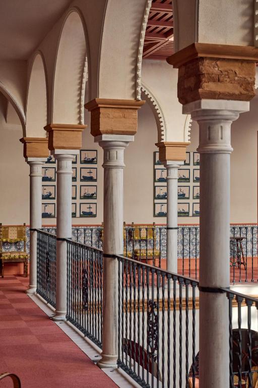 A corridor in the style of boutique hotels in Seville features arched columns with ornate capitals, overlooking a staircase. Framed art and blue patterned tiles adorn the walls, while a rich red carpet covers the floor.