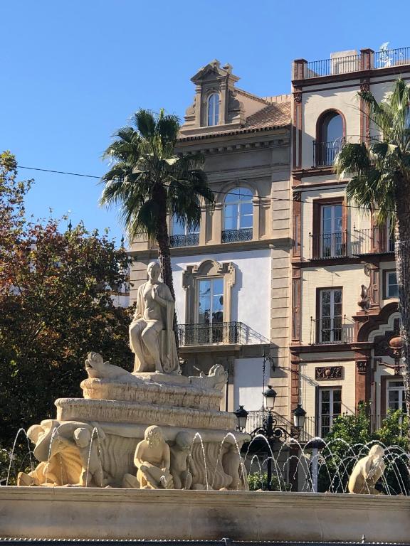 A stone fountain with lion sculptures and water streams, topped by a seated statue, is set in front of elegant historic buildings and palm trees under a clear blue sky—just steps away from some of the finest boutique hotels in Seville.