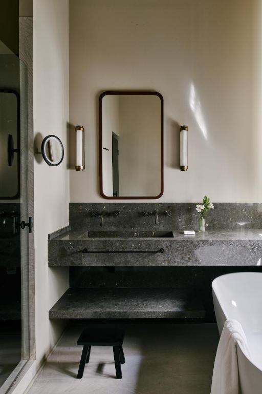 Modern bathroom with a large dark stone sink, wall-mounted mirror, two vertical lights, a small vase with flowers, a towel on a bathtub, and a black stool under the sink—evoking the chic elegance often found in boutique hotels in Seville.