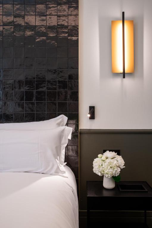 A neatly made bed with white linens beside a dark nightstand holding a vase of white flowers; reminiscent of the elegant comforts found in boutique hotels in Seville, a modern wall sconce emits warm light above.
