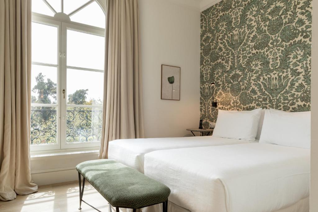 Bright bedroom with two white twin beds, a green cushioned bench, patterned green wallpaper, large window with beige curtains, and a framed picture on the wall—evoking the stylish charm often found in boutique hotels in Seville.