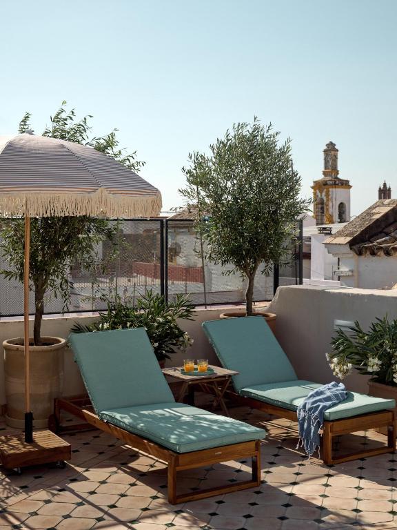 Two wooden lounge chairs with teal cushions sit on a tiled rooftop terrace at one of the boutique hotels in Seville, shaded by a fringed umbrella and surrounded by potted plants, with historic buildings visible in the background.