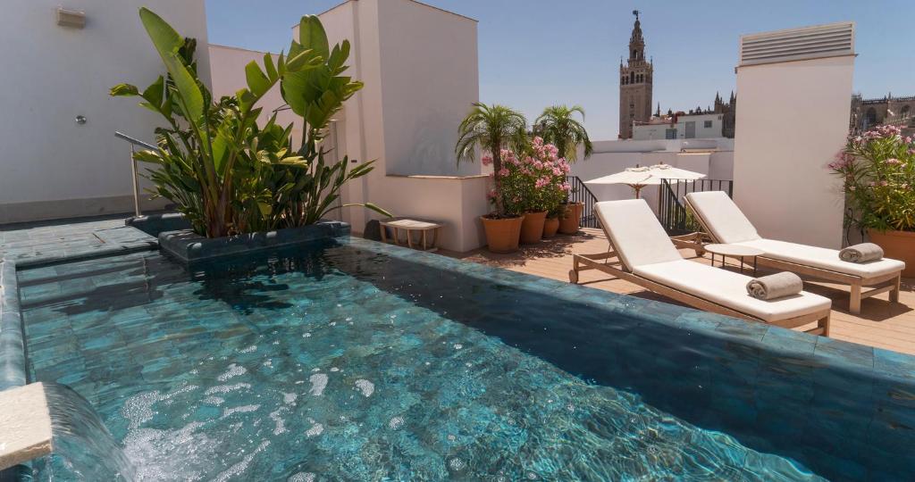 A rooftop pool with clear water, surrounded by potted plants and two white lounge chairs under a blue sky, offers the perfect retreat often found at boutique hotels in Seville. In the background, a historic tower rises above the cityscape.