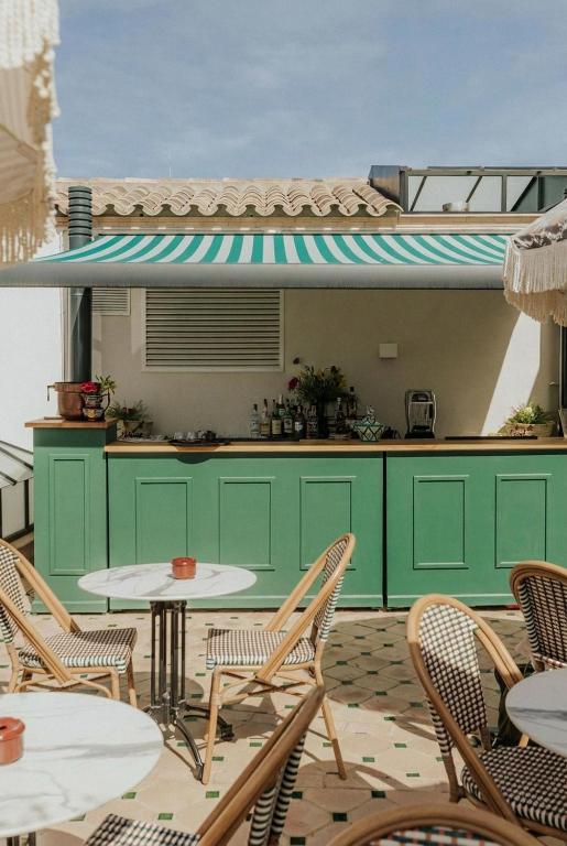 Outdoor bar area with green counter, striped green-and-white awning, and wicker chairs around small round tables. Sunlit setting with decorative tile floor and a relaxed, summery vibe typical of boutique hotels in Seville.