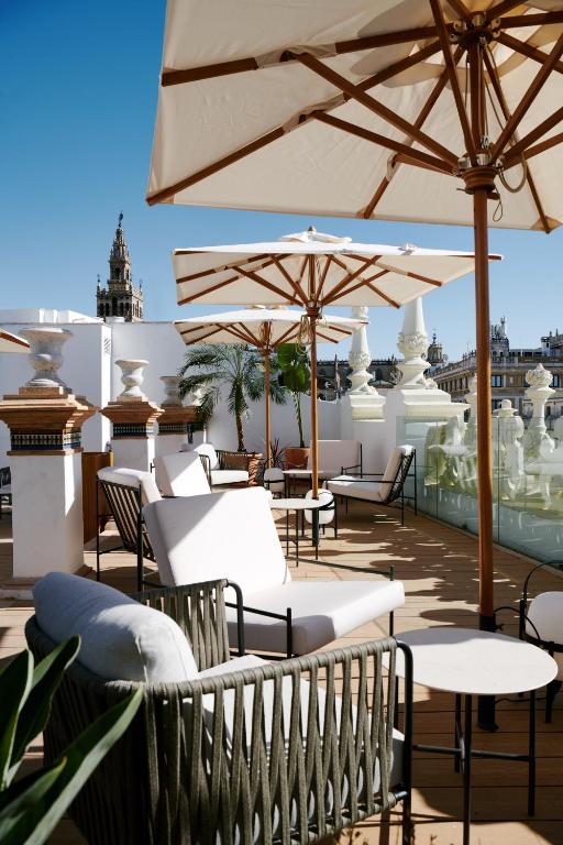 Outdoor rooftop terrace with cushioned chairs, tables, and large white umbrellas. Ornate white railings and city buildings are visible in the background under a clear blue sky, offering the kind of charming setting you’d expect from boutique hotels in Seville.