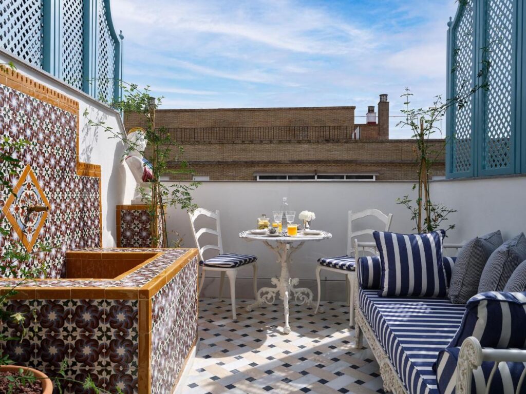 A cozy outdoor terrace, reminiscent of boutique hotels in Seville, features patterned tiles, a small round table set for drinks, striped cushions on a bench, and potted plants, all framed by blue lattice panels under a sunny sky.