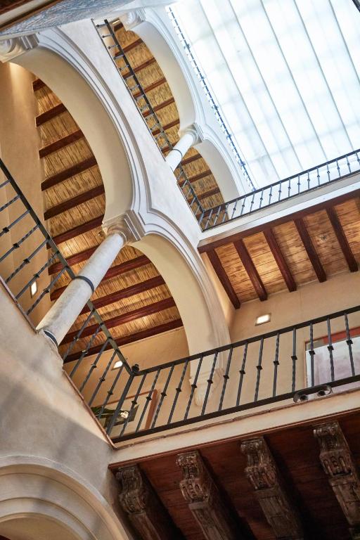 View looking up at multiple levels of a building with wooden ceilings, arched columns, and metal railings, reminiscent of the elegant interiors found in boutique hotels in Seville, with natural light coming through a large window above.