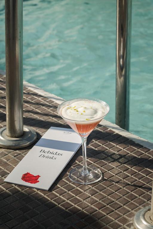 A pink cocktail in a martini glass sits on poolside tiles next to a drink menu, with metal pool railings and blue water in the background—a scene reminiscent of the chic ambiance found at boutique hotels in Seville.