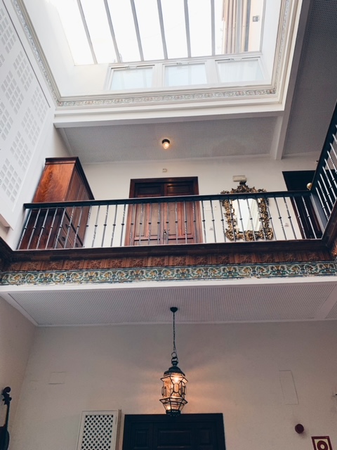 Reminiscent of the elegant charm found in boutique hotels in Seville, this two-story interior features a hanging lantern, wooden balcony railing, ornate framed mirror, and a large skylight that bathes the space in natural light from above.