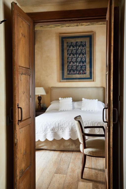 Wooden double doors open to a cozy bedroom reminiscent of the inviting charm found in boutique hotels in Seville, with a white bed, two pillows, a bedside lamp, a chair, and a framed artwork on the wall above the headboard. The floor is wooden, and the decor is rustic and warm.