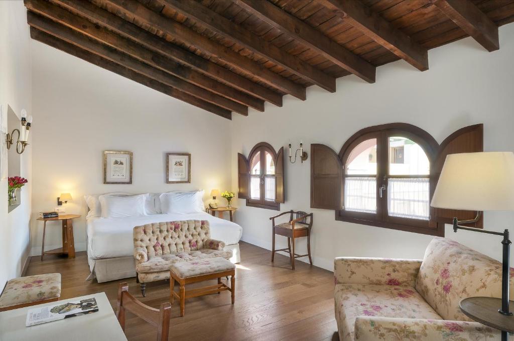 A cozy bedroom reminiscent of boutique hotels in Seville, featuring a wooden beamed ceiling, a double bed, floral sofa, armchair, desk, and arched windows with shutters. The room is bright with natural light and decorated in a charming rustic style.