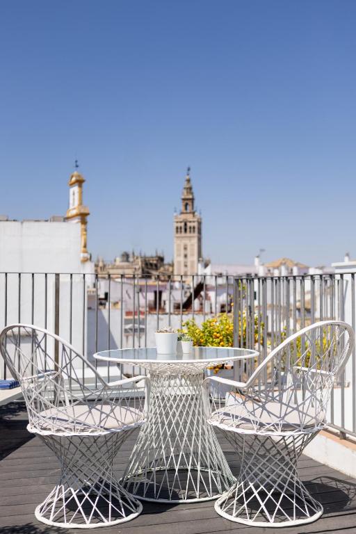 A rooftop terrace with two white wire chairs and a matching table, overlooking a cityscape with a tall historic tower under a clear blue sky—an inviting scene reminiscent of the stylish ambiance found at boutique hotels in Seville.