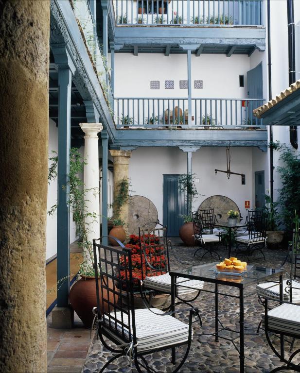 A charming courtyard reminiscent of the intimate spaces found in boutique hotels in Seville, featuring iron chairs and tables, potted plants, a cobblestone floor, and two stories of blue wooden balconies. Orange flowers and a bowl of lemons sit invitingly on the tables.