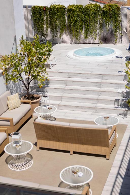 Modern outdoor patio with wicker furniture, round tables, and potted plants, reminiscent of the inviting style found at boutique hotels in Seville. Steps lead up to a small circular hot tub, surrounded by white tiles and green vines on a fence in the background.