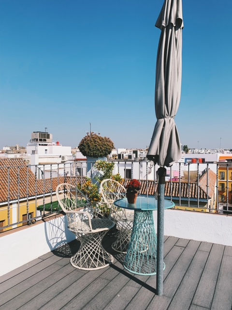 A rooftop patio inspired by the chic ambiance of boutique hotels in Seville, featuring wire chairs, a small glass table holding potted plants, and a closed gray umbrella. City buildings and clear blue sky form a stunning backdrop.