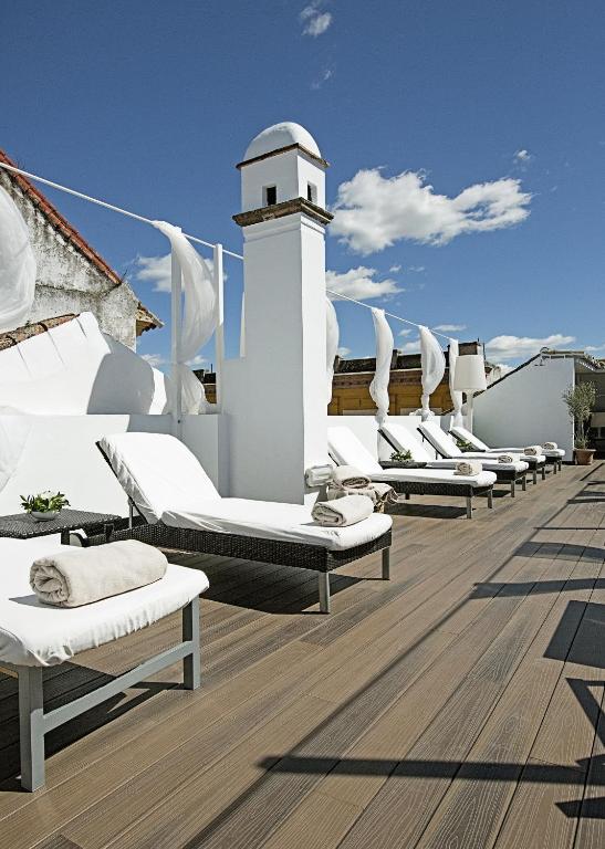 Several white lounge chairs with rolled towels sit on a wooden rooftop deck under a blue sky with clouds. White drapes and a white chimney add to the relaxing, luxurious atmosphere, reminiscent of the serene settings found at boutique hotels in Seville.